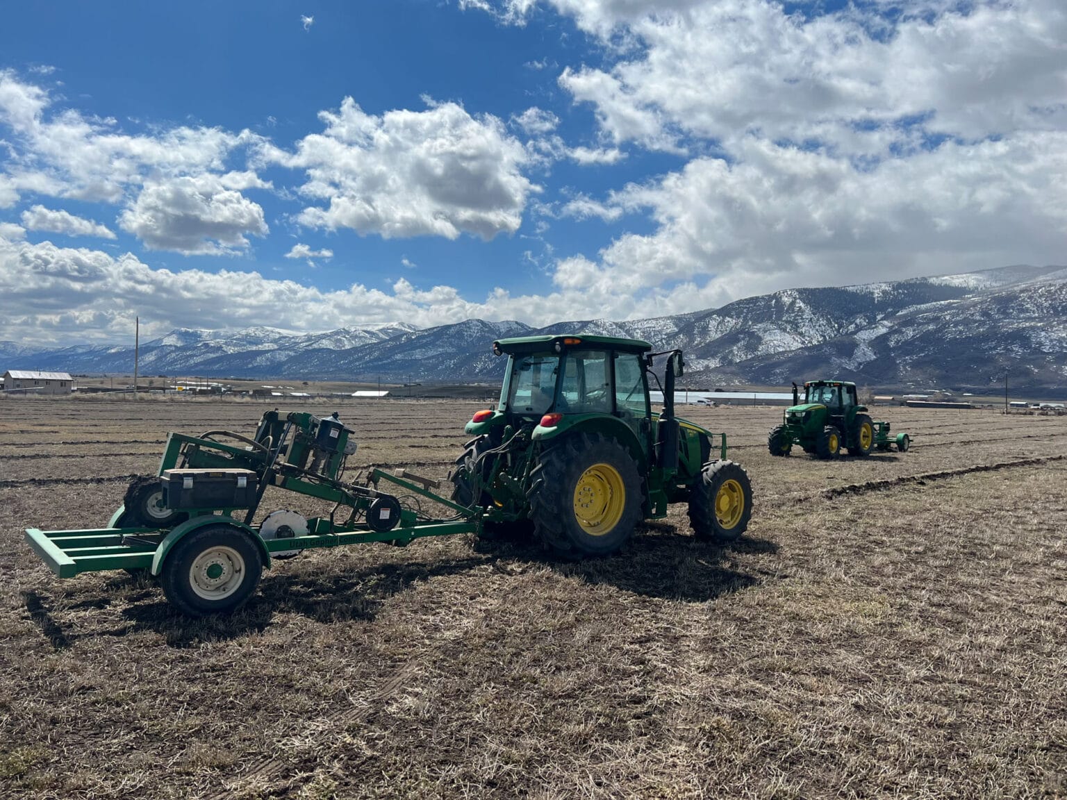 Two tractors working in field beneath snow-covered mountains