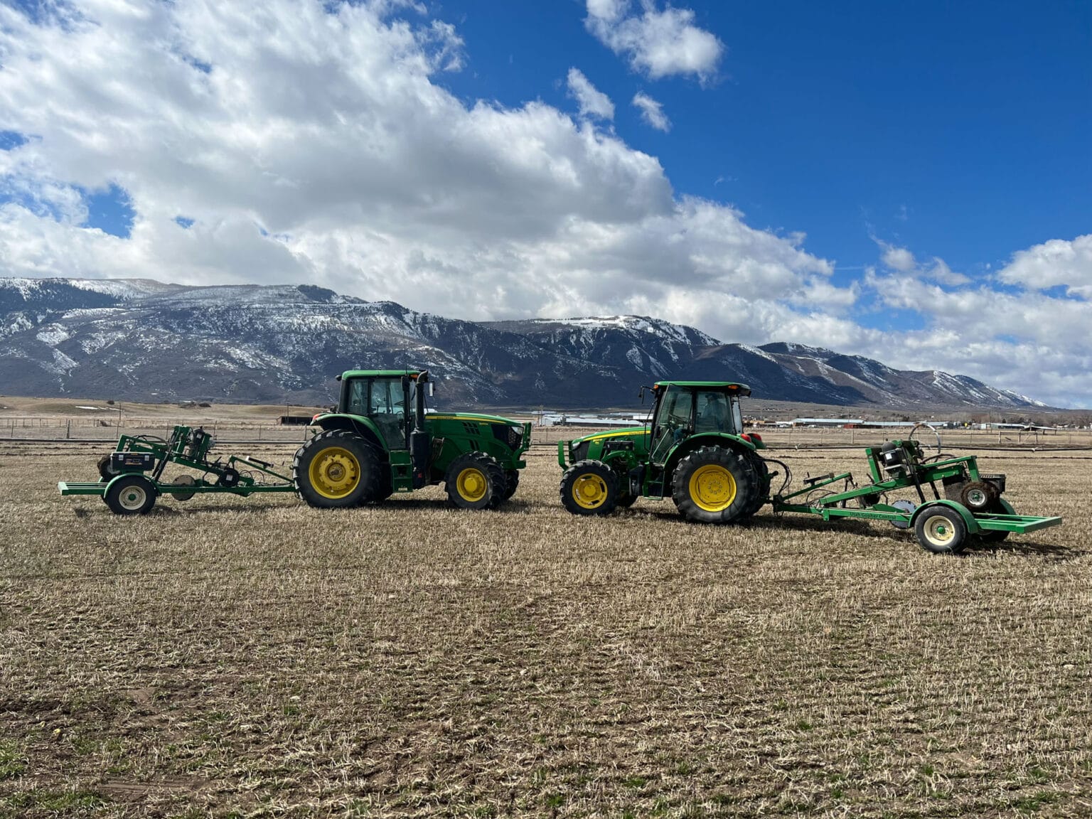 Two green tractors in field beneath snow-covered mountains
