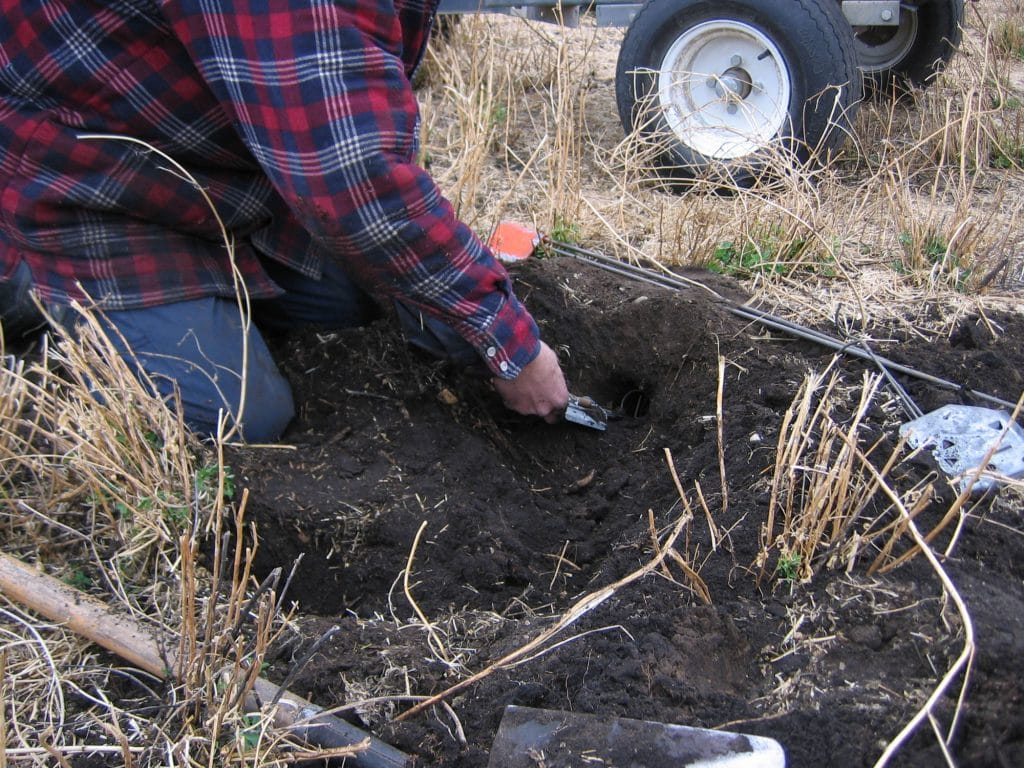 Person digging soil beside a wheeled cart in dry grass.