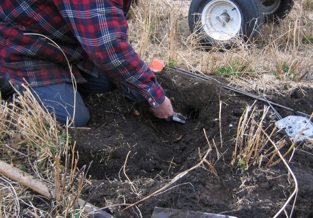 Person digging soil beside a wheeled cart in dry grass.